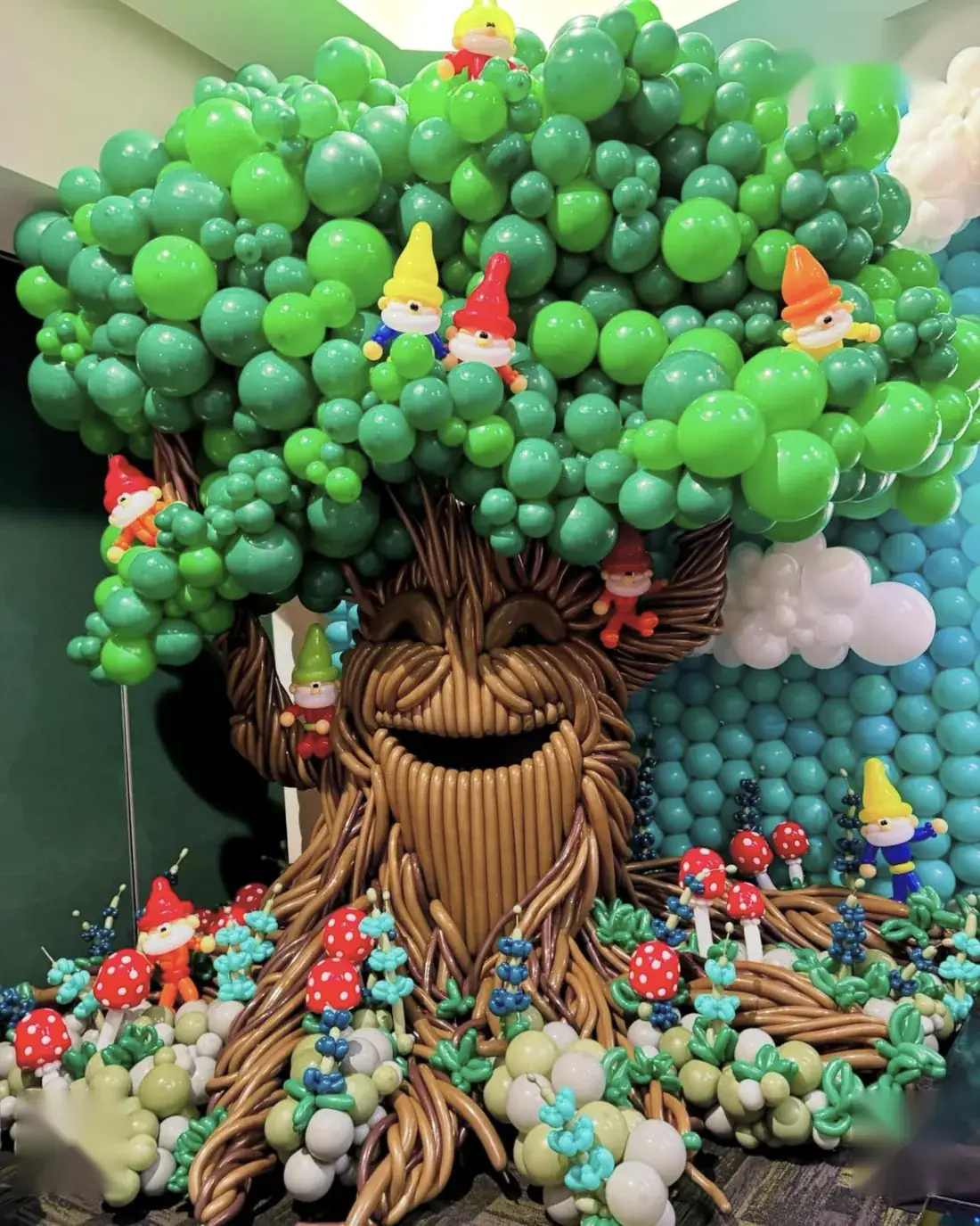 Árbol gigante de globos con cara sonriente, gnomos y hongos de globos