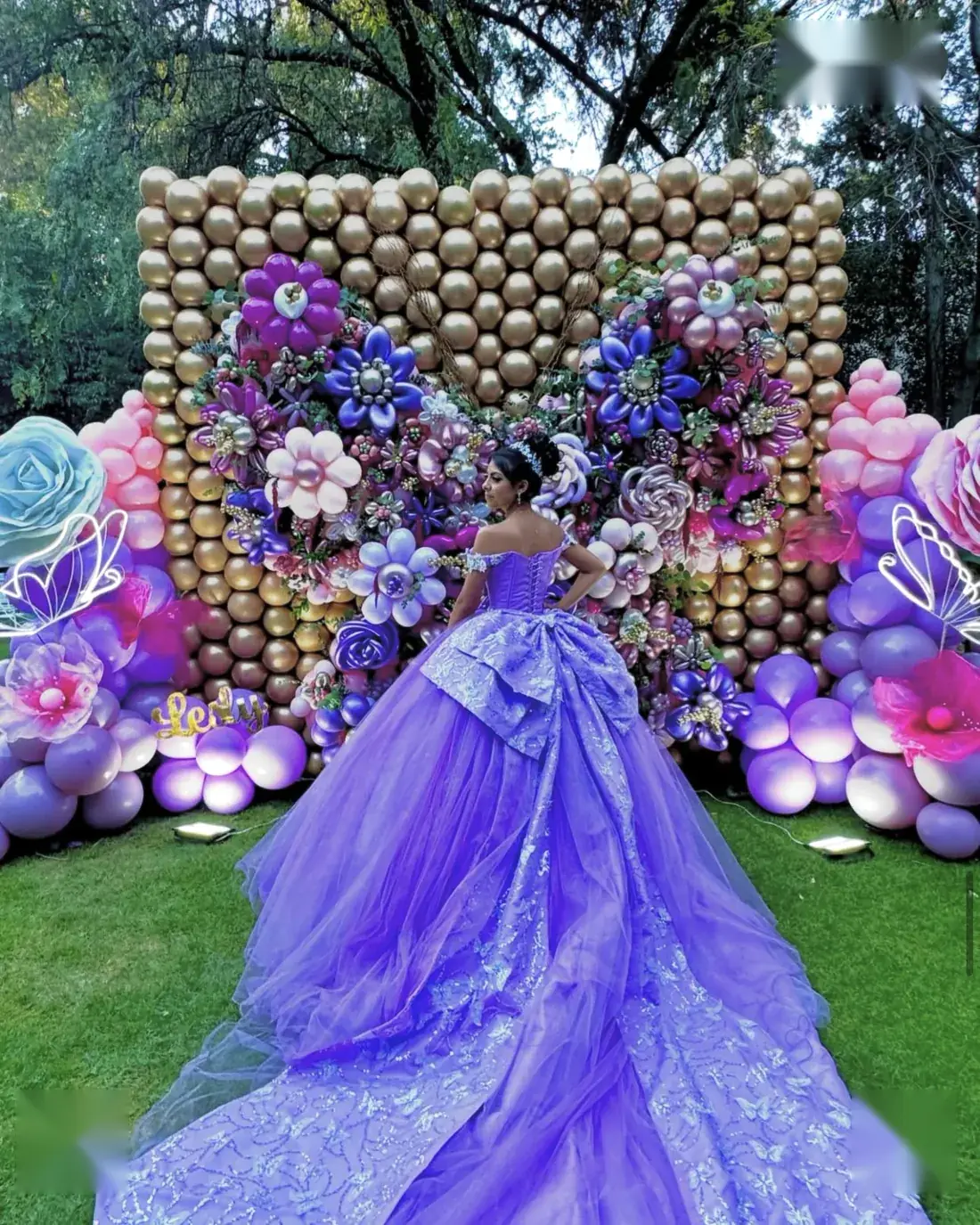 Quinceañera con vestido lila frente a un muro de globos dorados con flores gigantes
